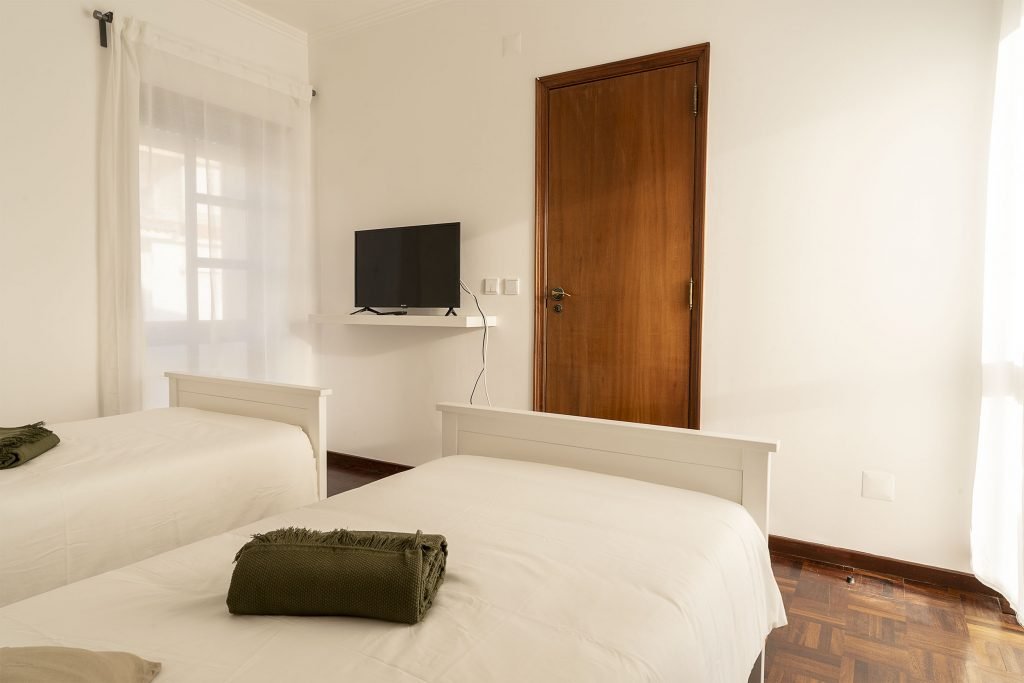 Two twin beds in a bright white room with neatly clean folded blankets and a TV